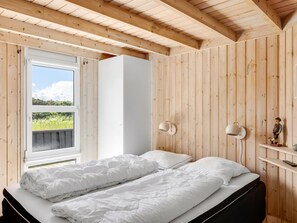 A bedroom with a wooden ceiling, a bed with white bedding, a window with a view of greenery, and two wall-mounted lamps.