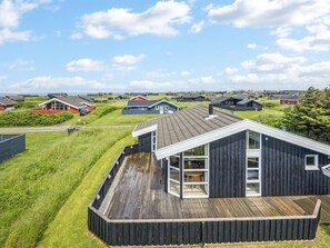 A modern house with a large deck, surrounded by a grassy area and other houses.