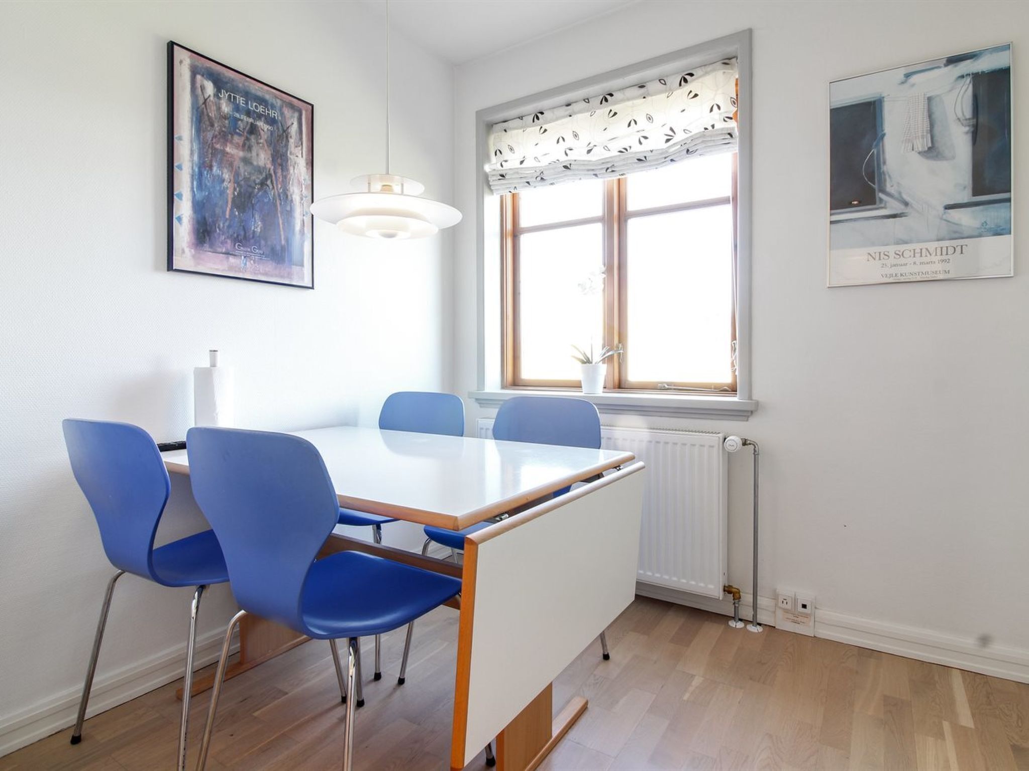 Un coin repas avec une table blanche et des chaises bleues, une fenêtre ornée de rideaux à motifs et des œuvres d’art accrochées aux murs.
