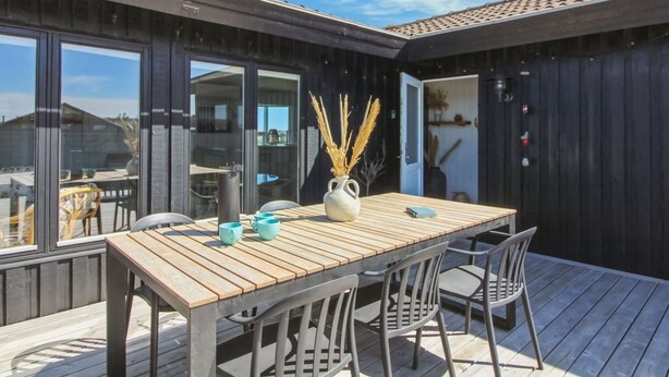 Una terraza de madera con mesa y sillas, ventanas amplias y vistas del entorno.