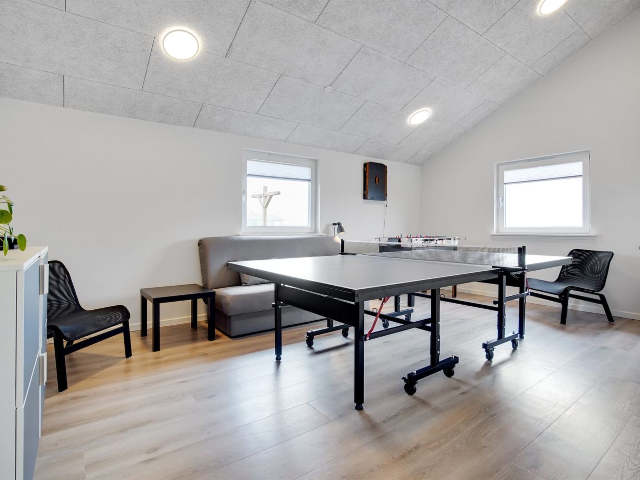 Una habitación moderna con mesa de ping-pong, dos sillas negras, un sofá gris y una mesita con una planta.