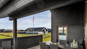 Un espace extérieur abrité, avec une table et des chaises, donnant sur des maisons au loin, et surmonté d’un mât de drapeau.