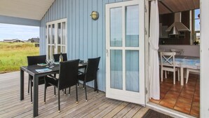 An open deck with a table and chairs, a sliding glass door leading to a kitchen, and a view of the surrounding landscape.