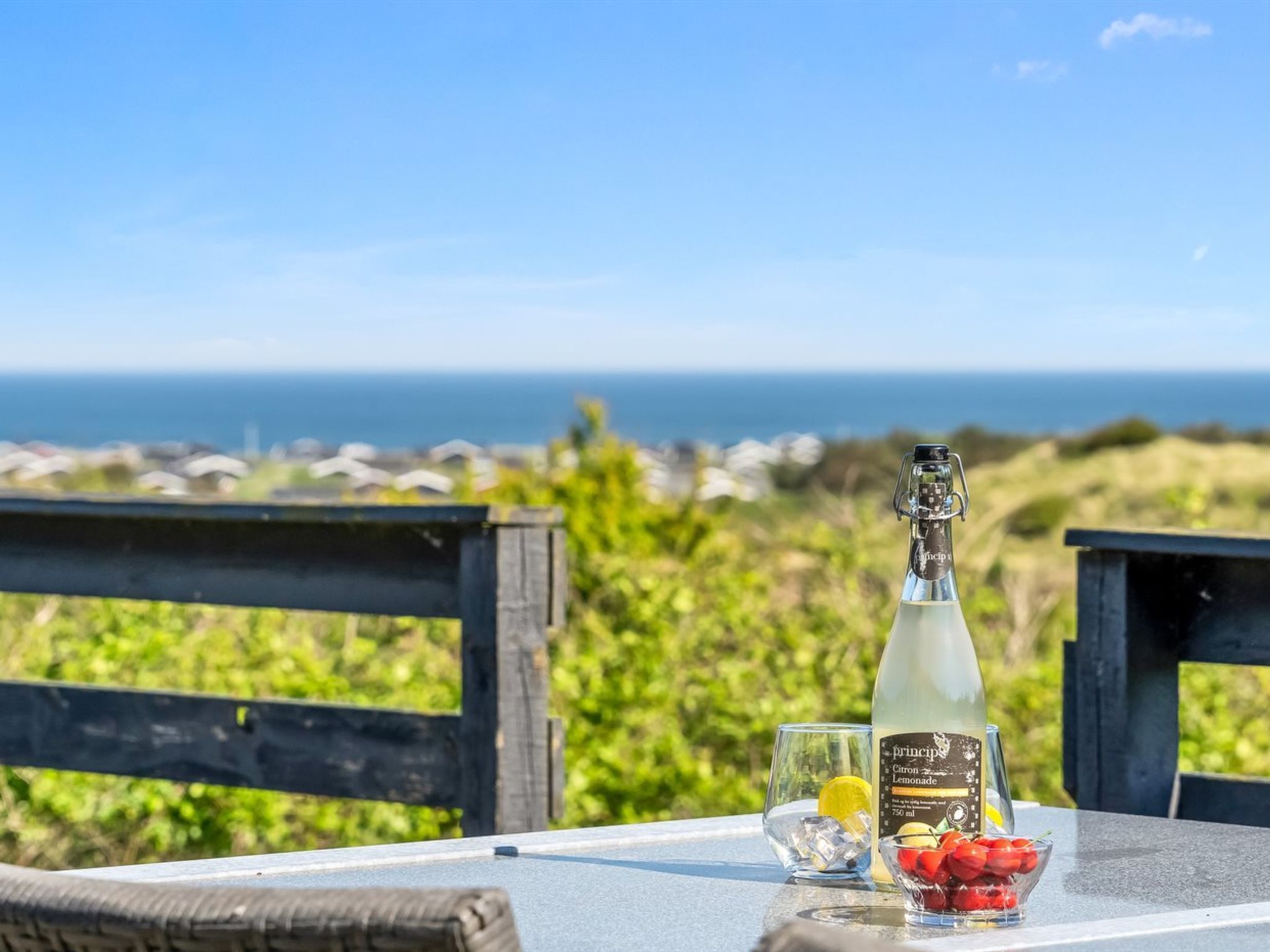 Ein Tisch mit einer Flasche Citron Zitronenlimonade, einem Glas mit Eiswürfeln und einer Schale Erdbeeren, vor einer Küstenlandschaft.
