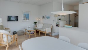 A modern living room with a white sofa, a glass coffee table, and wicker chairs. There is a dining table with chairs, a white pendant light, and a white ceiling.