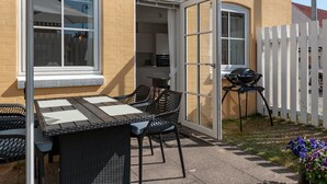 An outdoor patio with a table and chairs, a grill, and a white fence.