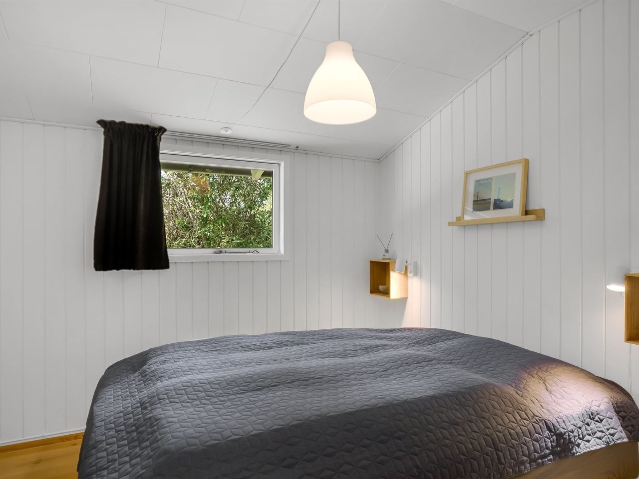 Une chambre à coucher avec un lit, une fenêtre donnant sur un paysage verdoyant, un luminaire suspendu et un tableau encadré au mur.