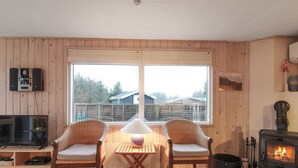 A cozy living room with wooden walls, a large window, a fireplace, two wicker chairs, a table with a lamp, and a TV on a wooden stand. - "Liisi" - 500m from the sea by Interhome (Hjørring)