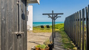 Ein Holzgebäude mit einer Metalttür, einer Beton-Terrasse mit Topfpflanzen, zwei Stühlen und Blick auf den Ozean.