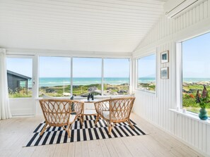 Een kamer met een groot raam dat uitkijkt op een strand, een geruit kleed en rieten stoelen.
