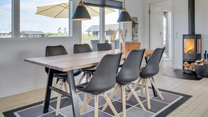 Un espace repas moderne avec une table en bois, des chaises noires et des luminaires suspendus. On aperçoit un poêle à bois à l’arrière-plan.