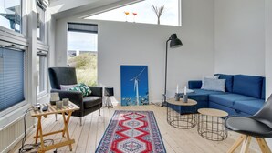 Un salon moderne avec un canapé bleu, un fauteuil noir, une table basse en bois, un tapis à motifs et un tableau représentant une éolienne accroché au mur.