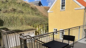 Ein Balkon mit zwei schwarzen Metallstühlen und einem kleinen Tisch, der auf ein gelbes Gebäude und einen Hügel mit Grünblick bietet.