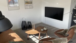 Ein modernes Wohnzimmer mit einem großen Flachbildfernseher, einer Couch, einem Couchtisch und zwei Sesseln. An der Wand hängen zwei gerahmte Bilder und auf dem Boden stehen zwei schwarze Taschen.
