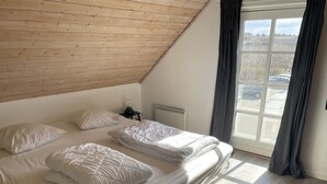 Ein Schlafzimmer mit schrägen Wänden, einem großen Fenster mit Vorhängen, einem Bett mit weißer Bettwäsche und einem kleinen Nachttisch mit Lampe.