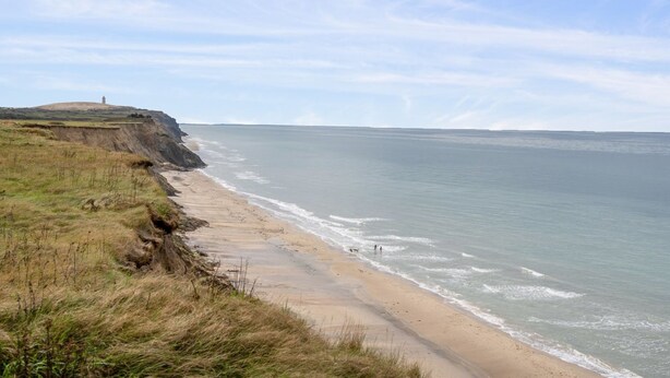 Eine Küstenlandschaft mit Sandstrand, Rasenflächen und einer Klippe mit einem Gebäude obenauf.