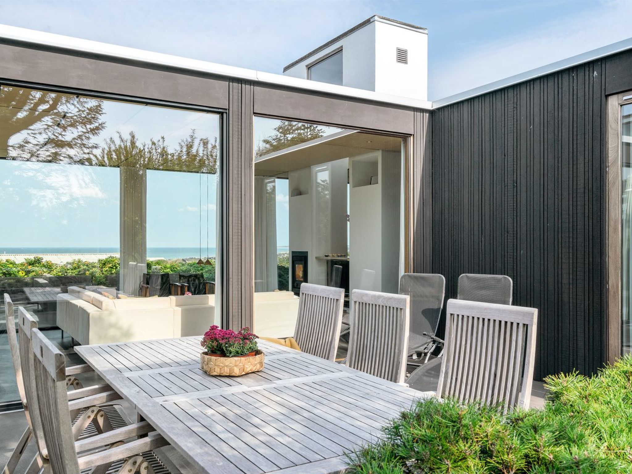 Un espace repas extérieur moderne, avec une table et des chaises en bois, une plante en pot et une vue sur l’océan.