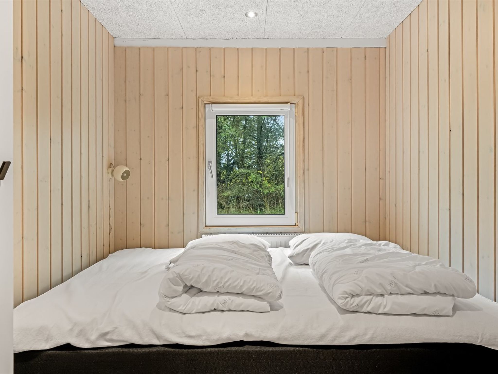 Quarto com parede de madeira, uma janela com vista para árvores e cama com roupa de cama branca.