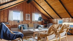 A cozy attic room with wooden walls and a sloped ceiling. There is a sofa, two chairs, a small table, and a bookshelf.