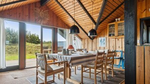 A dining area with a wooden table and chairs, large windows, and a view of the outdoors.
