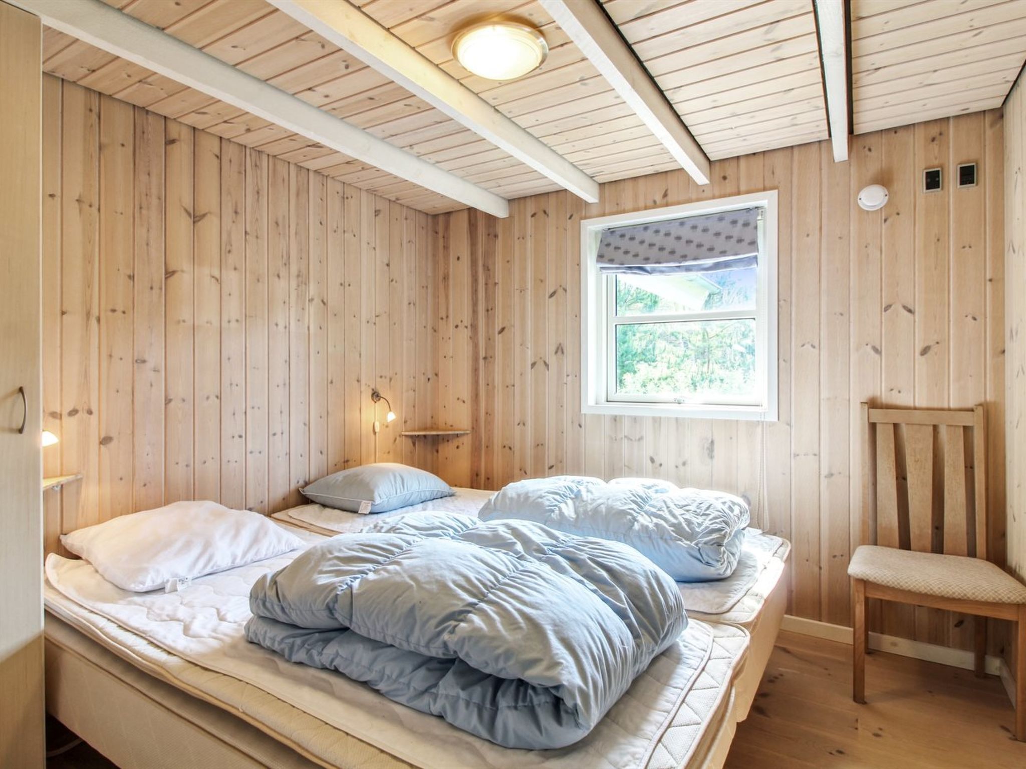 A wooden cabin room with a bed, a chair, and a window with a view of greenery.