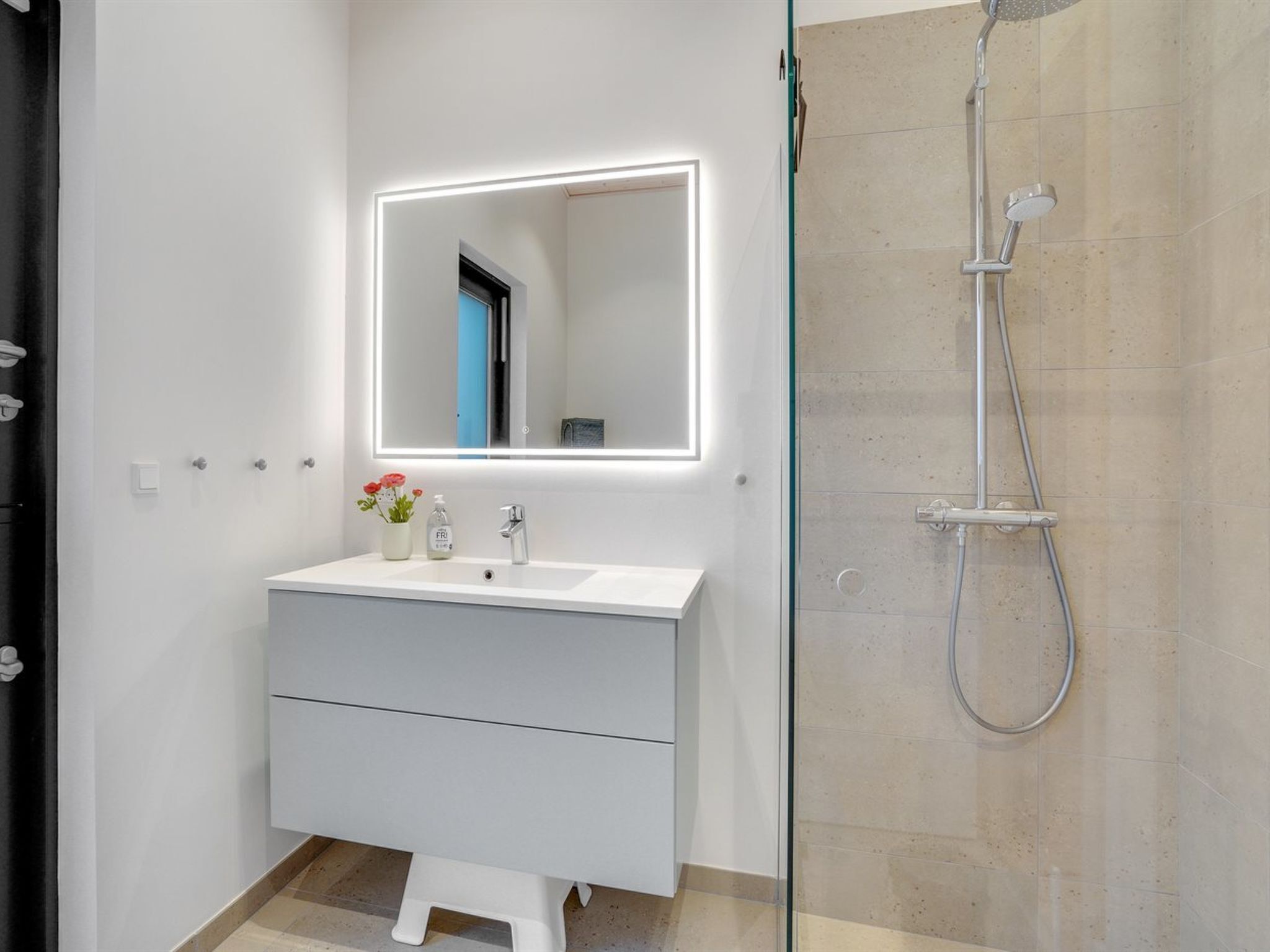 A modern bathroom with a white vanity, a mirror with LED lighting, a glass shower enclosure, and a small step stool.