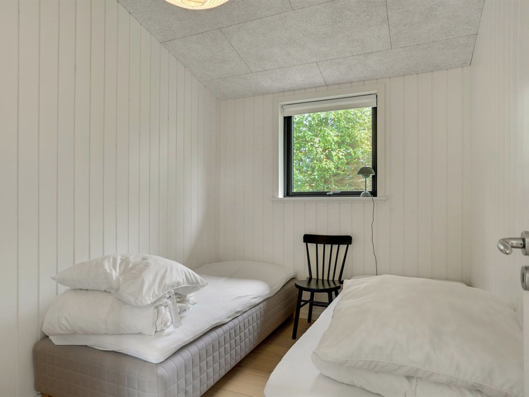 A small, single-room accommodation with a bed, a chair, and a window providing a view of greenery outside.