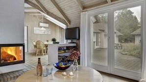 Une pièce chaleureuse avec une cheminée, une télévision, un coin repas avec des chaises et une grande fenêtre donnant sur un jardin.