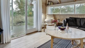 Une pièce chaleureuse avec un plafond en bois, une grande porte-fenêtre coulissante donnant sur un espace extérieur, une table ronde agrémentée d’un vase et de verres, et un canapé en cuir noir.
