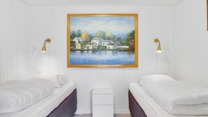 A bedroom with two beds, a painting of a lakeside village, and two wall-mounted lamps.