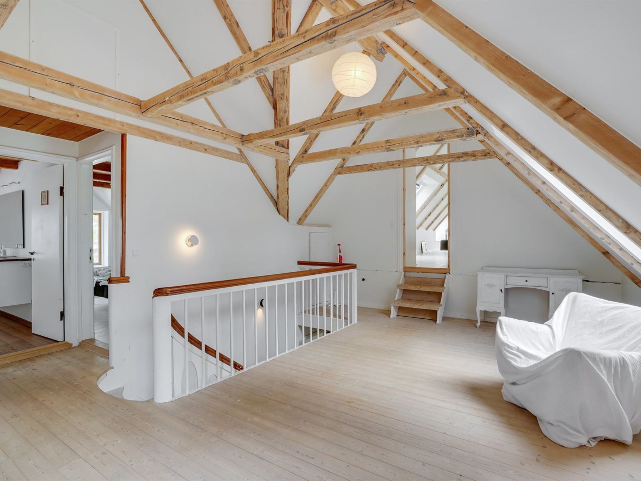 Een ruime zolderkamer met zichtbare houten balken, een witte trap en een groot raam voor natuurlijk licht.