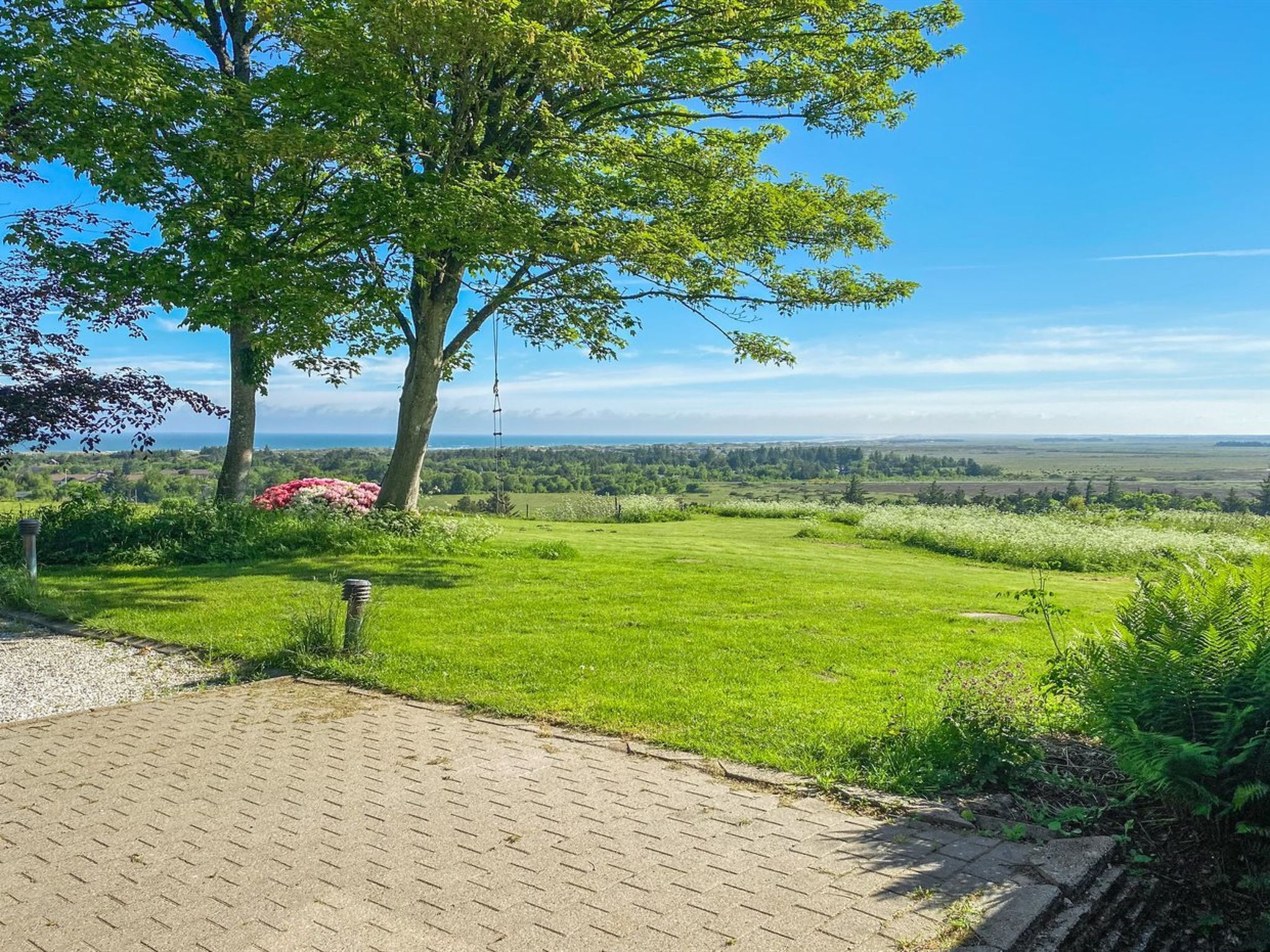 A paved pathway leading to a grassy area with a large tree and a clear view of the landscape.