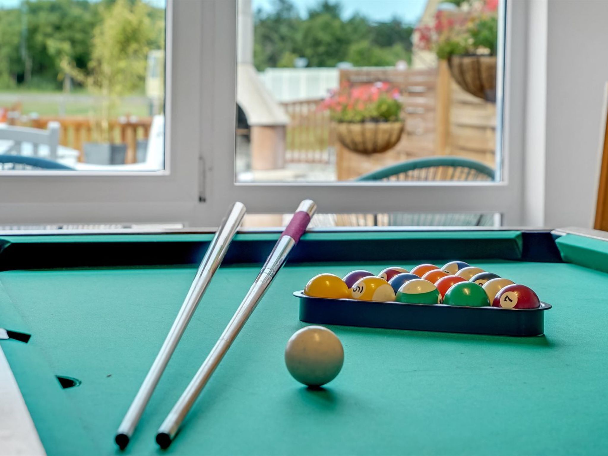 A pool table with balls and cues, a window with a view of outdoor seating and plants.