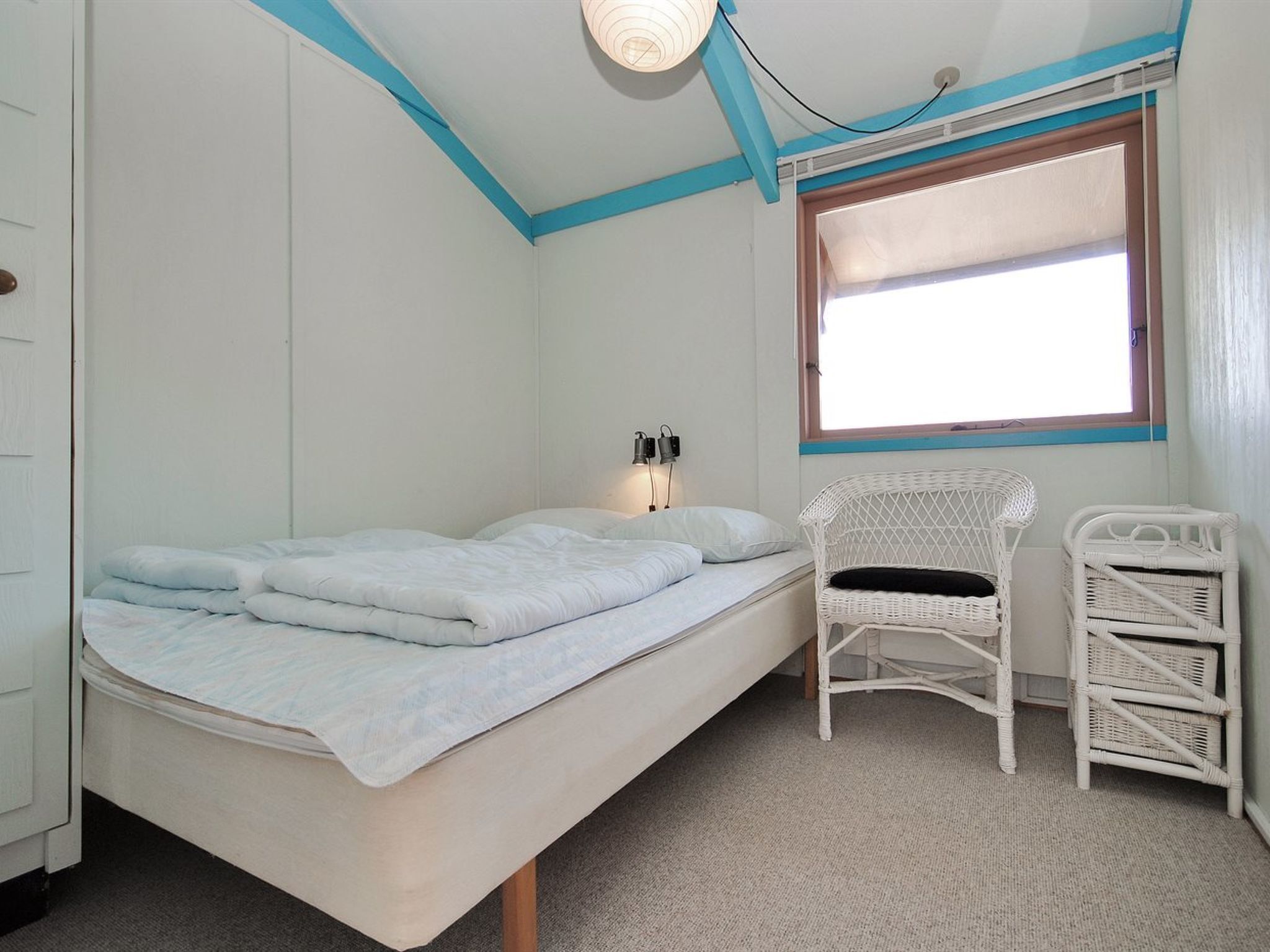 Una cama individual con colchón blanco y ropa de cama azul claro, una silla de mimbre blanca, una mesita pequeña y una ventana con marco de madera.