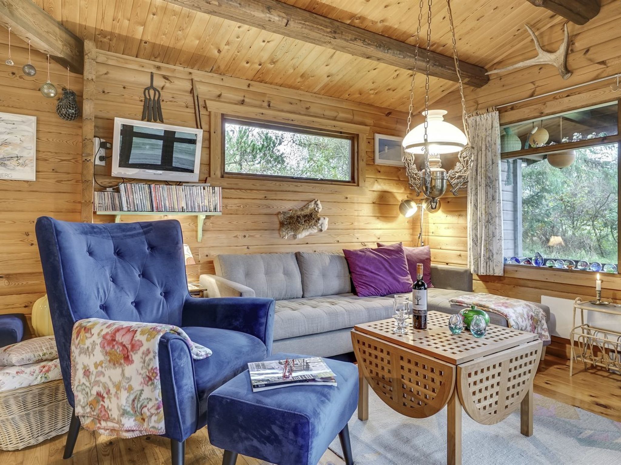 A cozy cabin interior with a blue armchair, a sofa, a small wooden table, and a television on the wall.