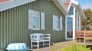A single-story building with a green exterior, white shutters, and a small porch area with a white bench and a blue plastic tub.