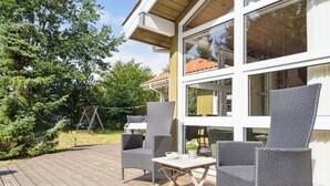 En träveranda med möbler av rotting och ett bord, med utsikt över en trädgård med träd och en gunga.