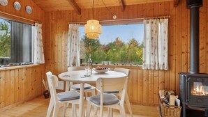 A cozy wooden cabin with a round dining table, four chairs, a hanging light fixture, a large window with curtains, and a wood-burning stove. - "Asmo" - 350m from the sea by Interhome (Fjerritslev)