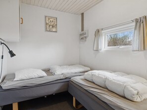 A dormitory room with two single beds, a desk, a lamp, a window with curtains, and a wall-mounted electrical outlet.