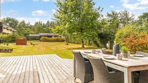 Een terras buiten met een houten tafel en rieten stoelen, omgeven door een gazon en bomen.