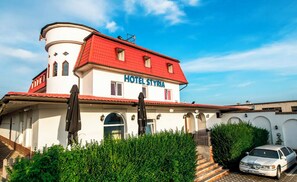 En tvåvåningsbyggnad med ett rött tak och vita väggar, med skylten ”HOTEL STYRIA”. Två svarta paraplyer står framför byggnaden, och en bil är parkerad på uppfarten.
