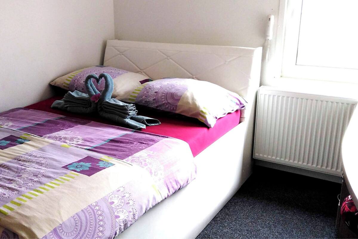 Una cama bien hecha con un colcha morada y blanca, una toalla doblada y una almohada con forma de corazón.
