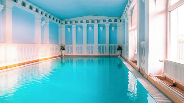 A large indoor swimming pool with clear blue water, surrounded by white columns and a high ceiling. - Hotel Styria (Chvalovice)