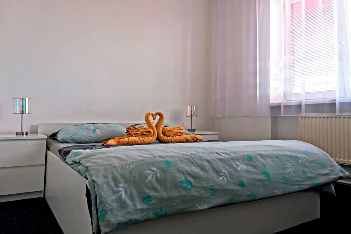 Una habitación de hotel con cama, dos mesitas de noche, ventana con cortinas translúcidas y un radiador.