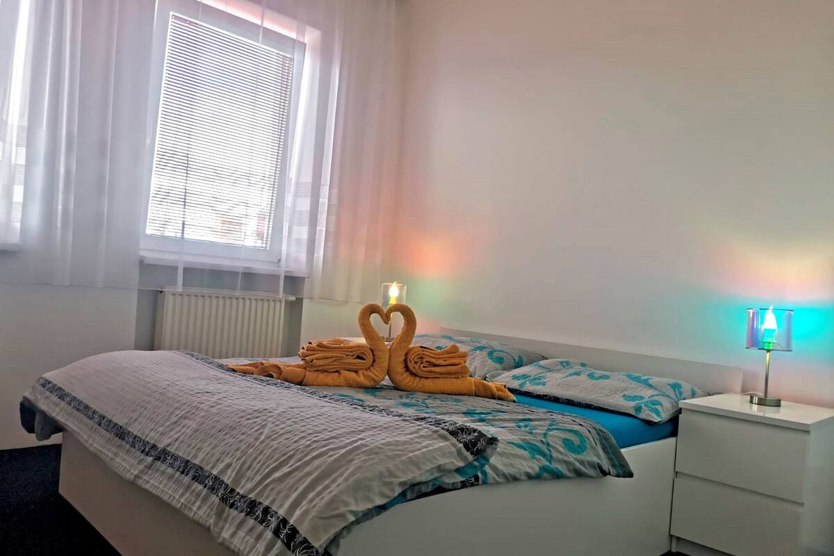 Una cama bien hecha con una colcha estampada, una lámpara de noche y una ventana con persianas.