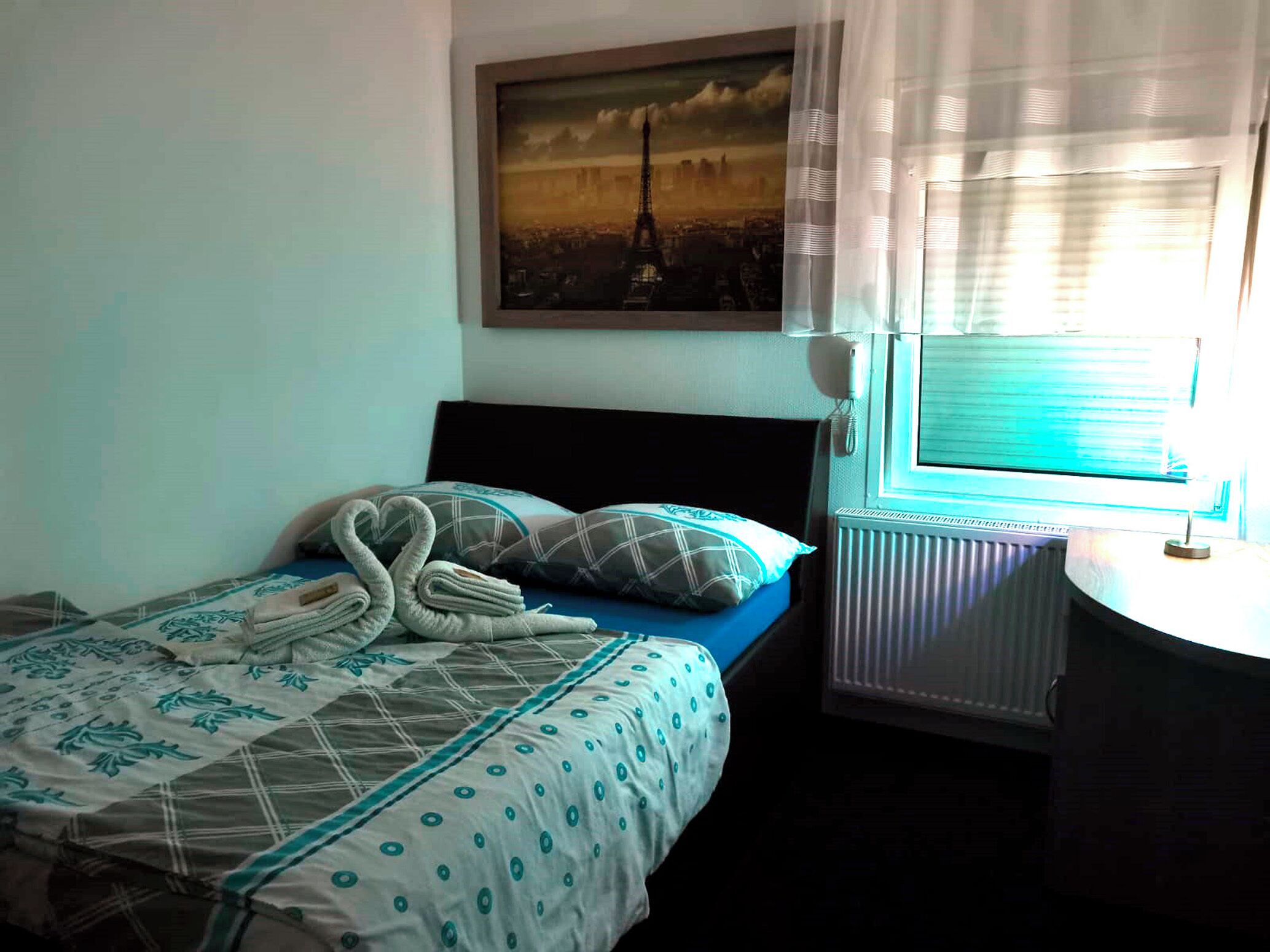 Una cama bien tendida con una colcha azul y blanca estampada, una toalla con forma de corazón y un cojín de diseño a cuadros.