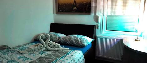 Una cama bien tendida con una colcha azul y blanca estampada, una toalla con forma de corazón y un cojín de diseño a cuadros.