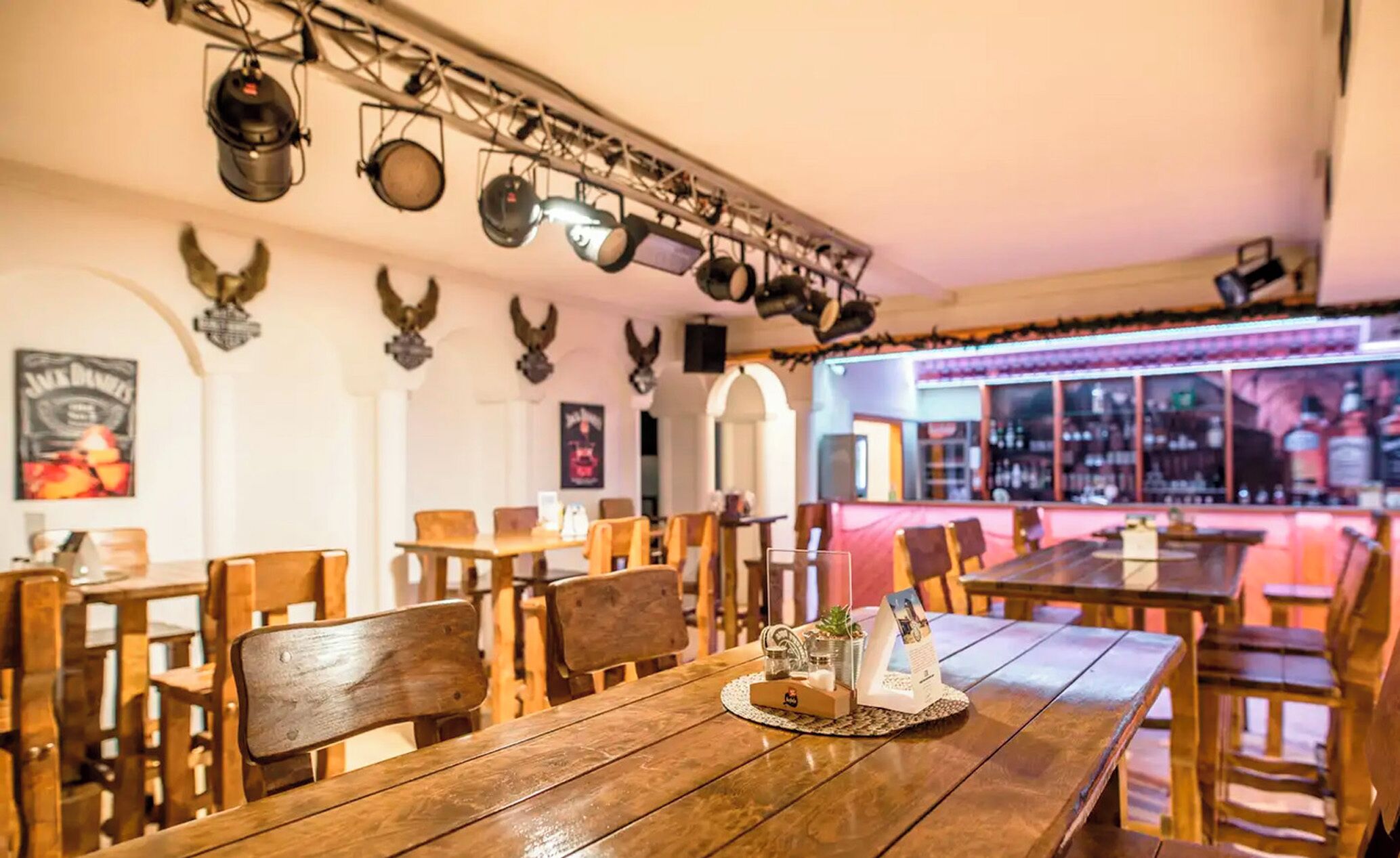 A restaurant with wooden tables and chairs, a bar area with bottles, and decorative wall-mounted lights.