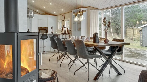 Une cuisine moderne équipée d’un poêle à bois, un coin repas avec une grande table et des chaises, et une vue sur l’extérieur grâce à de grandes fenêtres.