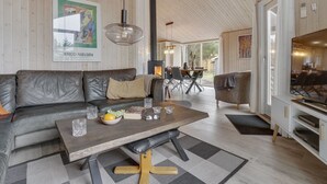 Un salon chaleureux avec un canapé en cuir, une table basse en bois et un coin repas comprenant une table et des chaises. Un grand tableau orne le mur, et une cheminée se trouve au fond de la pièce.
